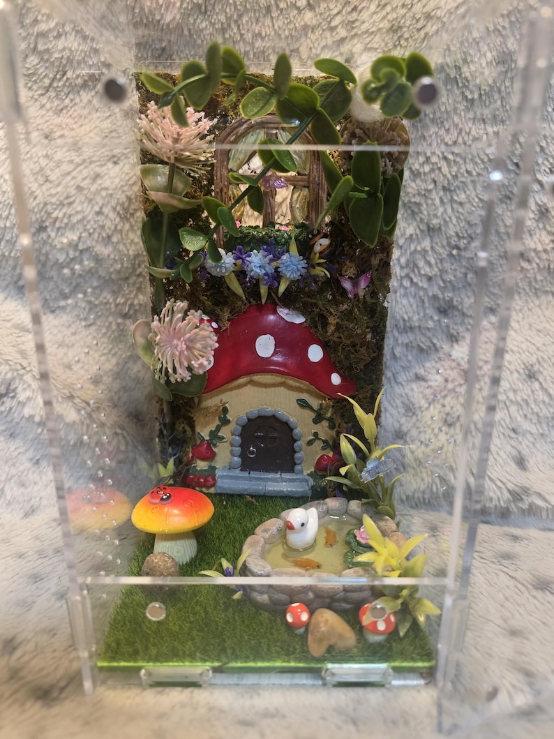 Puede incluir: Una escena de jard&iacute;n de hadas en miniatura encerrada en una caja de acr&iacute;lico transparente. La escena presenta una casa de hongos con una tapa roja y puntos blancos, un estanque con un pato blanco y varias plantas y flores.