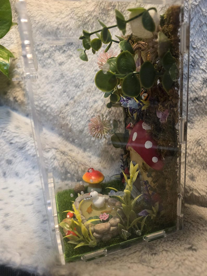 Puede incluir: Una escena de terrario en miniatura dentro de un recipiente de pl&aacute;stico transparente. La escena incluye un estanque con un pato, setas y varias plantas. Las setas tienen sombreros rojos con puntos blancos.