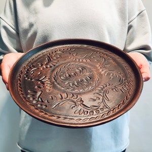 May include: A round, brown ceramic tray with an intricate, embossed floral design. The tray features a central floral motif surrounded by swirling patterns and leaf-like details. The edges are slightly raised, and the overall aesthetic is rustic and handcrafted.