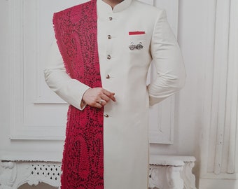 May include: A white long-sleeved kurta with a mandarin collar and button closure. The kurta has a red embroidered shawl draped over the left shoulder. The man is wearing white pants.