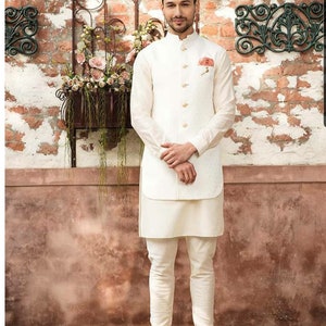 Puede incluir: Un hombre con una kurta blanca y un chaleco blanco con un patr&oacute;n floral. Est&aacute; de pie frente a una pared de ladrillo con una maceta colgando.