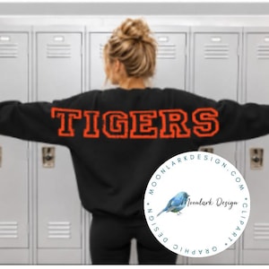 May include: A black long-sleeve sweatshirt featuring the word TIGERS printed in large, bold, orange block letters across the back. The garment has a crew neckline and a relaxed fit, shown from the back against a background of gray lockers.