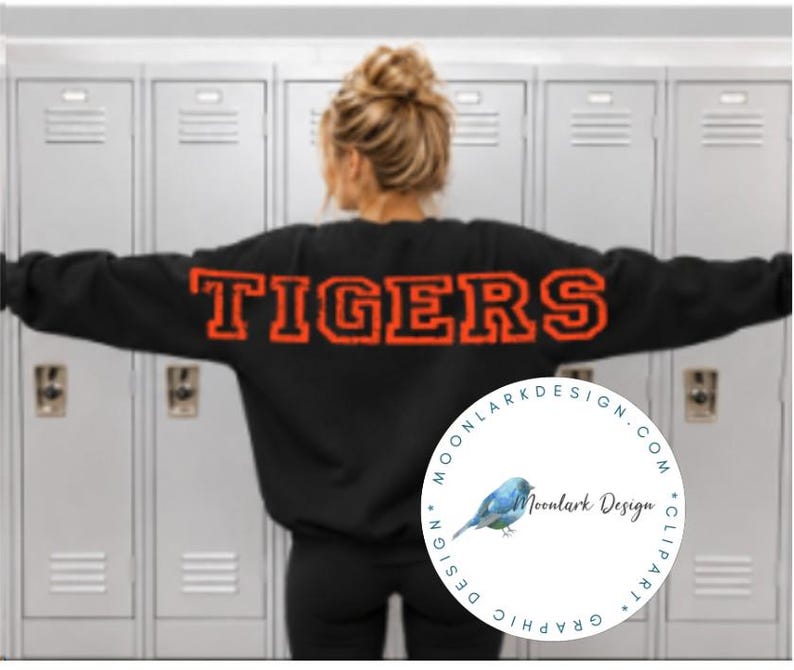 May include: A black long-sleeve sweatshirt featuring the word TIGERS printed in large, bold, orange block letters across the back. The garment has a crew neckline and a relaxed fit, shown from the back against a background of gray lockers.