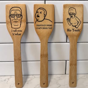 May include: Three light brown bamboo kitchen spatulas featuring laser-engraved illustrations and quotes. The left spatula shows a man with glasses and the text I tell you h'what. The center shows a boy and the text THAT'S MY PURSE I DON'T KNOW YOU. The right shows a woman and the text Ho Yeah!