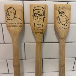 May include: Three wooden cooking spoons with engraved cartoon characters and phrases. One spoon features Bobby Hill with the text "THAT'S MY PURSE I DON'T KNOW YOU." Another has Hank Hill with "I tell you h'what." The third spoon has Peggy Hill with "Ho Yeah!"