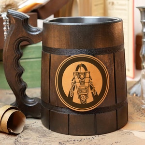 Puede incluir: Un jarro de cerveza de madera marrón oscuro con un asa decorativa. El jarro presenta un emblema circular con un diseño de robot negro y dorado. El jarro tiene un interior de acero inoxidable y mide aproximadamente 15 cm de alto.