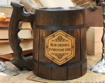 May include: A dark wooden beer mug with a stainless steel interior and a large handle. The mug has a leather patch with the text "Rob Drinks, Everyone Dies". Two silver candlesticks flank the mug.