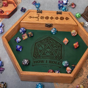 May include: A hexagonal wooden dice tray with a green felt lining featuring a printed 20-sided die graphic and the text "HOW I ROLL". The tray includes a hinged wooden lid with engraved dice icons. Scattered polyhedral dice surround the tray.