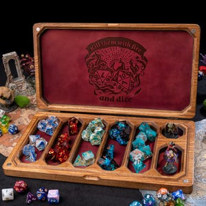 May include: Wooden dice box with a red velvet interior, featuring the text "Kill them with fire and dice." The box is open, revealing various colorful polyhedral dice. Miniature figurines and terrain pieces surround the box.