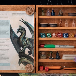 May include: Wooden tabletop game organizer with a hinged door. The left side features a printed page with a dragon illustration and text. The right side has shelves holding dice, miniature figures, and small bottles. A green Sharpie marker is visible.