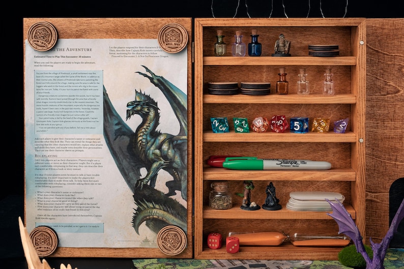 May include: Wooden tabletop game organizer with a hinged door. The left side features a printed page with a dragon illustration and text. The right side has shelves holding dice, miniature figures, and small bottles. A green Sharpie marker is visible.