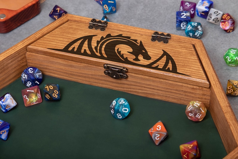 May include: A wooden box with a green felt interior and a dragon design on the lid. The box is filled with colorful dice, including a blue d20, a red d6, and a green d12.