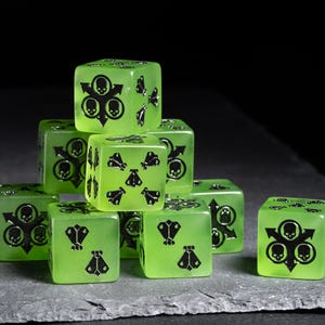 May include: A collection of translucent green dice with black symbols, including skulls and arrows. The dice are stacked on a gray surface against a black background. The dice are a standard size, approximately 0.75 inches per side.