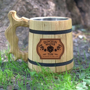 May include: A wooden beer stein with a stainless steel interior. The stein has a black band around the top and bottom and a brown leather patch with the text "Dungeon Master Tom The Weaver of Lore & Fate" and a 20-sided die.