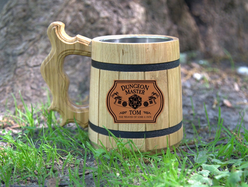 May include: A wooden beer stein with a stainless steel interior. The stein has a black band around the top and bottom and a brown leather patch with the text "Dungeon Master Tom The Weaver of Lore & Fate" and a 20-sided die.