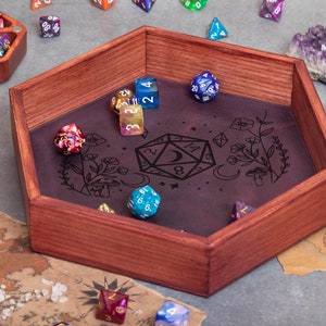 Puede incluir: Una bandeja hexagonal de madera para dados con un forro de fieltro morado. La bandeja tiene un diseño de un dado D20 con una luna creciente y flores. Hay varios dados de colores dentro de la bandeja.