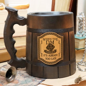 Peut inclure: Une chope en bois brun foncé avec une sangle en cuir noir et un couvercle en métal. La chope a un patch en cuir brun avec le texte "When the DM smiles it's already too late" et un graphique d'un dé.
