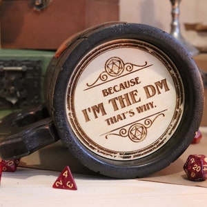 May include: A black and brown ceramic mug with a wooden handle. The mug has a white circle on the front with the text "Because I'm the DM That's why." and a stylized image of a 20-sided die. The mug is surrounded by red dice.