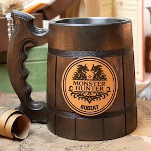 Puede incluir: Un jarro de cerveza de madera marrón oscuro con un asa decorativa. El jarro presenta un emblema circular con las palabras "Monster Hunter" y el nombre "Robert". El interior del jarro es de metal.