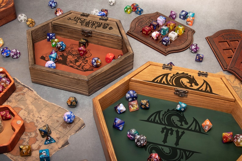 May include: Two wooden dice boxes with engraved designs. One box is hexagonal and has a goblin design with the text "Dice Goblin Vault". The other box is rectangular and has a dragon design with the text "TY". Both boxes are open and filled with colorful dice.