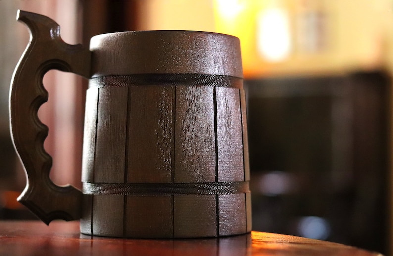 Pode incluir: Uma caneca de madeira castanho-escura com um design semelhante a um barril, com ripas verticais e duas faixas horizontais. A caneca tem uma pega grande e curva com um padr&atilde;o de ader&ecirc;ncia entalhado distinto e acabamento polido.