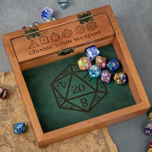 May include: A square wooden dice box with a hinged lid featuring the engraved text "choose your weapon" and various dice shapes. The interior base is lined with dark green material displaying a large 20-sided die graphic, containing several multi-coloured polyhedral dice.