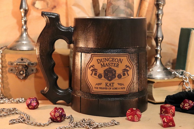 May include: A brown wooden beer stein with a black handle and a leather label that reads "Dungeon Master Kat The Weaver of Lore & Fate". The label features a 20-sided die with the numbers 20 and 9 visible. There are several red dice on a table in front of the stein.