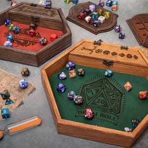 May include: Two hexagonal wooden dice trays with hinged lids and metal latches. One tray features a red felt interior with the text The Dice Giveth and the Dice Taketh Away, while the other has a green felt interior with a large d20 graphic and the text Roll.