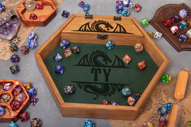 May include: A wooden hexagonal dice tray with a green felt interior. The tray has a dragon design and the name "TY" engraved on it. The tray is filled with colorful dice.