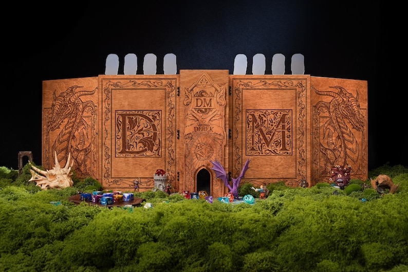May include: A wooden DM screen with dragon and letter engravings. The screen is brown with a central tower design and the letters "DM" on the front. Miniature figures and dice are arranged on a bed of green moss.