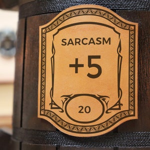 Może przedstawiać: Brązowy drewniany kubek z sk&oacute;rzanym naszywką, na kt&oacute;rej widnieje napis "SARCASM +5 20".