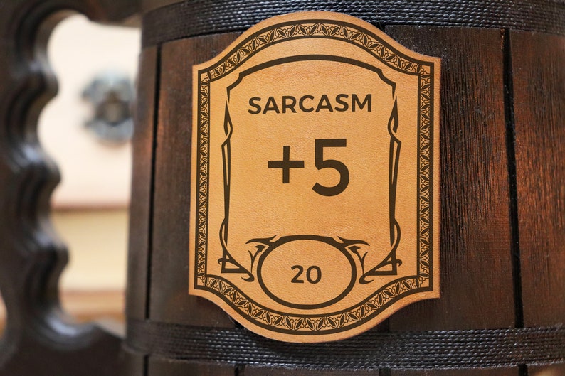 Może przedstawiać: Brązowy drewniany kubek z sk&oacute;rzanym naszywką, na kt&oacute;rej widnieje napis "SARCASM +5 20".
