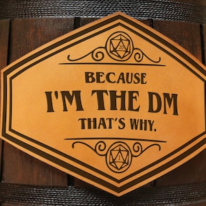 Pode incluir: Um barril de madeira castanho-escuro com uma placa hexagonal de material semelhante a couro bege com o texto preto "BECAUSE I'M THE DM THAT'S WHY." A placa inclui desenhos de linhas pretas de um dado de vinte lados no topo e um dado de um lado na base.