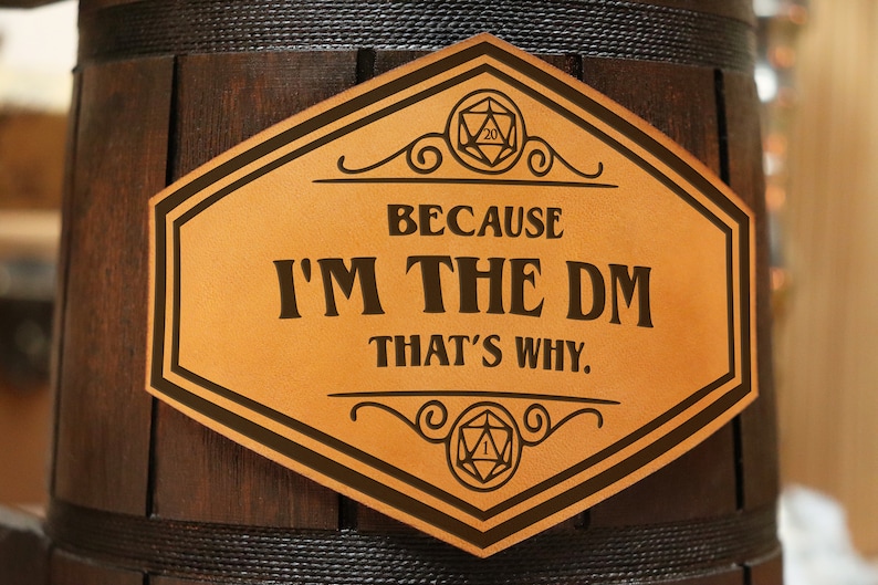 Pode incluir: Um barril de madeira castanho-escuro com uma placa hexagonal de material semelhante a couro bege com o texto preto "BECAUSE I'M THE DM THAT'S WHY." A placa inclui desenhos de linhas pretas de um dado de vinte lados no topo e um dado de um lado na base.