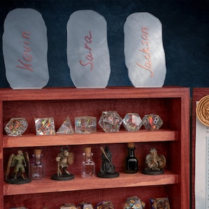 May include: A wooden display case with shelves holding various gaming dice, miniature figurines, and small glass bottles. Above the case are three frosted glass nameplates with names written in red. A wooden coin with a logo is on the right.