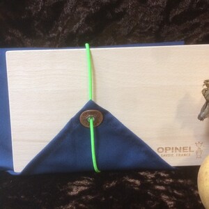 May include: A wooden cutting board with a blue fabric cover and a green elastic band. The board has the text "Opinel Savoie France" printed on it.