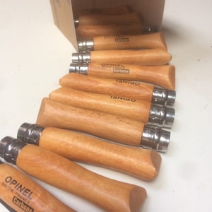 May include: A row of ten wooden-handled pocket knives with stainless steel blades. The knives are all the same size and have the word "Opinel" engraved on the handle. The knives are arranged in a line on a white surface.
