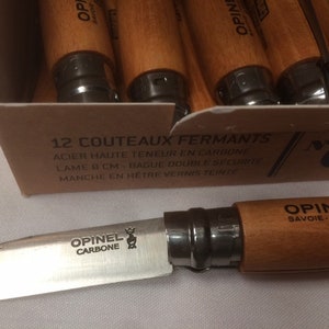 May include: A box of 12 Opinel folding knives with carbon steel blades and beechwood handles. The box is labeled "12 Couteaux Fermants" and "No. 02".