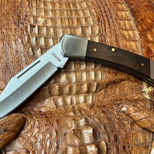 以下が含まれることがあります： 茶色のハンドルと銀色の刃を持つポケットナイフ。刃が開いており、ナイフは茶色のワニ革の表面に置かれています。ナイフには刃に「WHITBY」という文字が刻まれています。