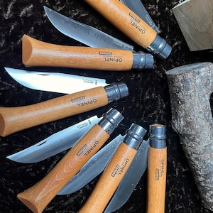 May include: A collection of eight Opinel folding knives with wooden handles and stainless steel blades. The knives are arranged in a circle on a dark brown surface. The knives are all different sizes and have the Opinel logo and "Carbone" printed on the blades.
