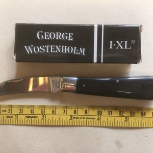 May include: A black handled pocket knife with a silver blade. The knife is closed and is sitting on a yellow measuring tape. The knife is labeled "I-XL" and "GEORGE WOSTENHOLM".