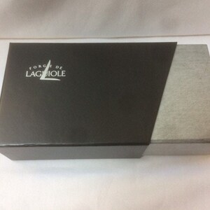May include: A black box with a white logo that says "Forge de Laguiole" on the top. The box is made of cardboard and has a gray fabric lining.