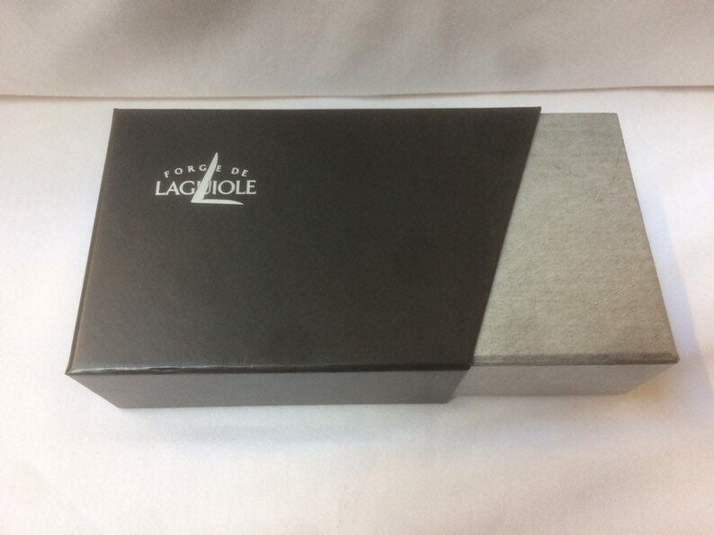 May include: A black box with a white logo that says "Forge de Laguiole" on the top. The box is made of cardboard and has a gray fabric lining.