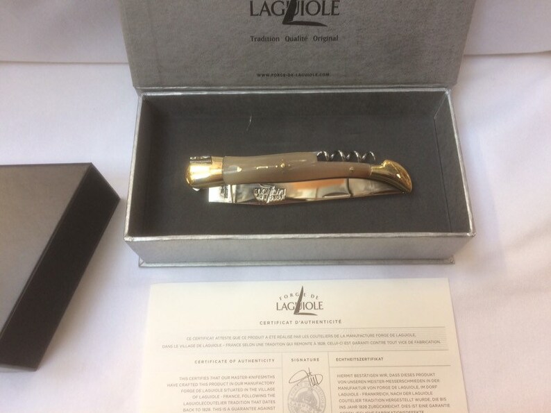 May include: A Laguiole pocket knife with a bone handle and gold accents, in a gray box with a certificate of authenticity. The certificate states that the knife was handcrafted in the village of Laguiole, France.