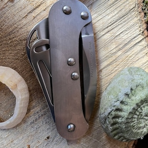 May include: A closed, silver-colored pocket knife with a curved handle and a partially visible blade. The knife is attached to a white rope and has the text "Reg. Des. 300724" printed on the side. The knife is resting on a wooden surface.