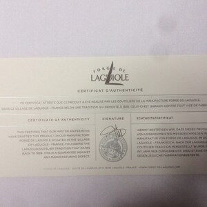May include: A white certificate of authenticity for a product made by Forge de Laguiole. The certificate features the company logo, a signature, and text in French, English, and German. The text states that the product was made in the village of Laguiole, France, following a tradition that dates back to 1828. The certificate guarantees the product against any manufacturing defects.