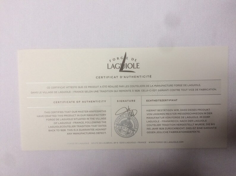 May include: A white certificate of authenticity for a product made by Forge de Laguiole. The certificate features the company logo, a signature, and text in French, English, and German. The text states that the product was made in the village of Laguiole, France, following a tradition that dates back to 1828. The certificate guarantees the product against any manufacturing defects.