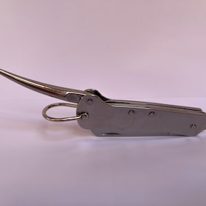 Puede incluir: Una multiherramienta de acero inoxidable con una hoja curva y puntiaguda y un pequeño lazo redondeado. La herramienta está cerrada, revelando un cuerpo rectangular compacto con remaches. La hoja está extendida, mostrando su diseño afilado y curvo.