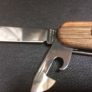 Könnte beinhalten: Ein silbernes Multifunktions-Taschenmesser mit einem Holzgriff. Der Griff hat eine Holzmaserung und das Victorinox Swiss Made Logo ist in den Griff graviert. Das Messer hat eine Klinge, einen Flaschenöffner und einen Dosenöffner.