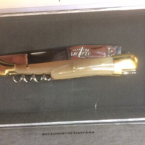 May include: A gold and ivory-colored pocket knife with a corkscrew and the words "Forge de Laguiole" on the blade. The knife is in a box with the website address "www.forge-de-laguiole.com".